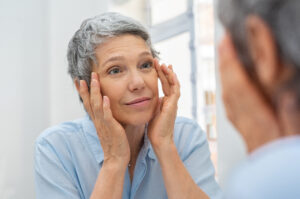 Mature woman looking in mirror and pulling up skin, considering a face lift