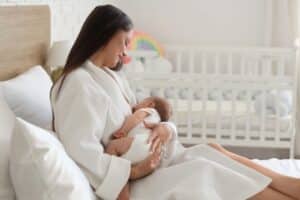 Happy young woman in a bathrobe breastfeeding her baby at home, showing successful lactation after breast reduction surgery.