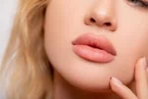 A close-up of a young woman’s perfectly plump lips in peach matte lipstick showcases the results of lip augmentation and expert makeup application.