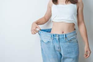Young, slim woman in oversized jeans, showing toned waist after weight loss from diet, exercise, and liposuction.